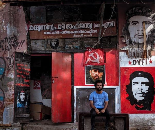 A man sits outside a building with communist motifs and writing in English and Malayalam, flags, and images of Che Guevara